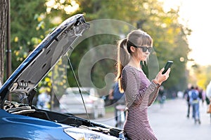 Young upset woman driver talking on mobile phone near a broken car with open hood waiting for help having trouble with her vehicle