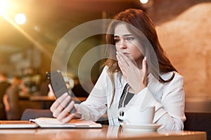 Young upset woman with phone in her hand working outside office in laptop looks shocked recieving bad news drinking hot coffee in