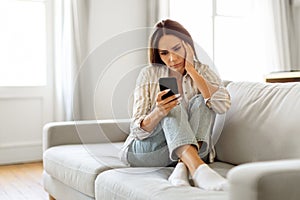 Young upset woman sitting on couch and looking at her smartphone