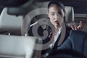 Young upset woman in suit sitting in backseat of car with digital tablet