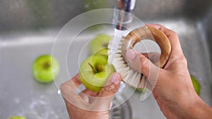 Young vegan girl washing green apples with bamboo brush. Hand holding fresh fruits under running water in kitchen sink