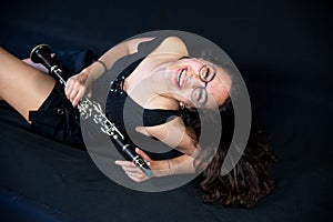 A brunette girl, with glasses, lying laughing and holding her clarinet music instrument isolated on a black background