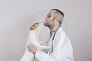 Young veterinarian man examining a cute small dog by using stethoscope, isolated on white background. Indoors. Dog kissing the