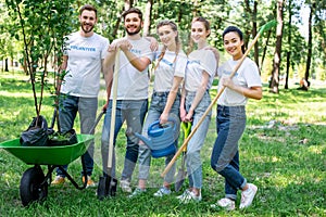 young volunteers posing in park with watering can shovel rake and wheelbarrow