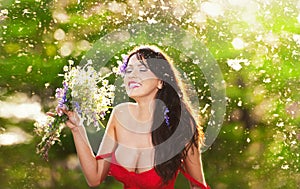 Young voluptuous brunette holding a wild flowers bouquet in a sunny day. Portrait of beautiful woman with low-cut red dress laugh