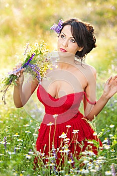 Young voluptuous brunette holding a wild flowers bouquet in a sunny day. Portrait of beautiful woman with low-cut red dress posing