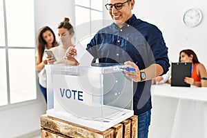 Young voter man smiling happy putting vote in voting box standing by ballot at electoral center