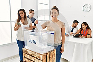 Young voter woman smiling happy putting vote in voting box standing by ballot at electoral center