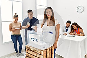 Young voter woman smiling happy putting vote in voting box standing by ballot at electoral center