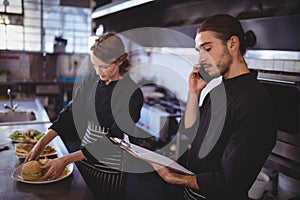 Young waiter talking on smartphone while waitress preparing food in commercial kitchen