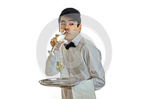 A young waiter in a white shirt with a butterfly holds a tray and drinking champagne from a glass