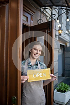 Young waitress standing at the door, holding open sign, smiling