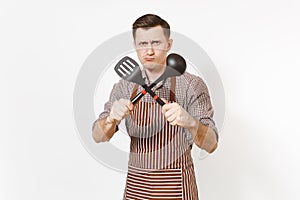 Young warlike man chef or waiter in striped brown apron, shirt holding black ladle or kitchen spoon, spatula isolated on