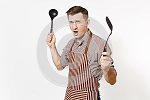 Young warlike man chef or waiter in striped brown apron, shirt holding black ladle or kitchen spoon, spatula isolated on