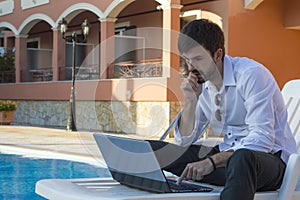 Young warried businessman working on his laptop by the pool whi