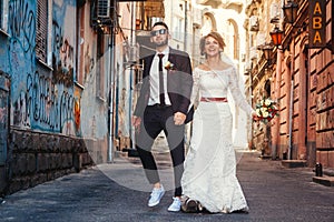 Young wedding couple celebrating marriage in city. Fun bride and groom smiling and walking in on modern city`s street.