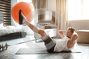 Young well-built man go in for sports in apartment. Holding big red fitness ball between feet and doing abs workout
