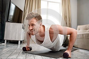 Young well-built man go in for sports in apartment. Strong muscular guy stand in plank position using dumbbells and look