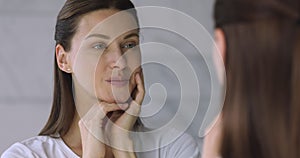 Young well-groomed lady touching face, looking at mirror.