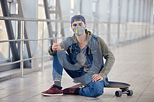 Young white man in hoody sitting on a skateboard on the street with a medical face mask on. Coronavirus concept photo