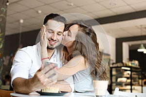 Young woman kissing man using smartphone at cafe and eating cake.