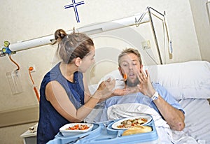 Young wife trying to feed his reluctant husband lying in bed at hospital room ill after suffering accident