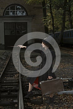 Young witch with magic wand conjures at old train station. Girl dressed in black cape, pointed hat and holds retro suitcase in