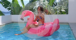 Young woman 25s in bikini using smartphone while resting at vacation travel, swimming on trendy inflatable pink flamingo