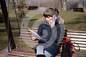 young woman accountant sits on a park bench and reads a reporting document and calls on a smartphone, reporting period