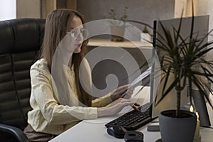 Young woman accountant works at computer and holds documents. Boss is in own office. Secretary enters data into program