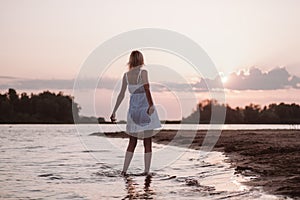 A young woman admires the sunset. A beautiful slender happy blonde in a white summer dress stands on the beach in the