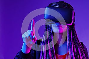 Young woman with afro braids using VR glasses on dark purple neon background