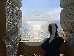 A young woman alone standing from the top of an old stone bell tower looking out at the vast ocean.