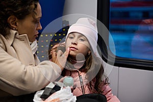 Young woman applies moisturizing balm on daughter lips, taking care of skin in the winter time while travelling by train