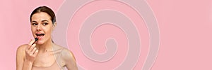 A young woman applies nude lipstick on her lips on a pink background. Cosmetics concept. Web banner