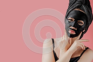 Young woman apply black cosmetic fabric facial mask on pink background. Face peeling mask with charcoal, spa beauty treatment,