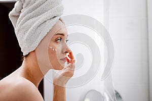 Young woman applying anti-wrinkle cream standing behind mirror in home bathroom. Cosmetology and beauty procedure. Skin care after