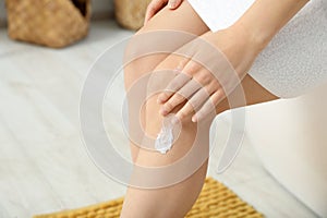 Young woman applying body cream on leg indoors, closeup
