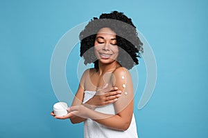 Young woman applying body cream onto shoulder on light blue background