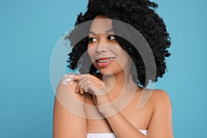 Young woman applying body cream onto shoulder on light blue background