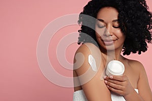 Young woman applying body cream onto shoulder on pink background. Space for text