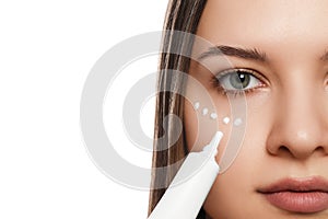 Young woman applying cream under eye on white background, closeup