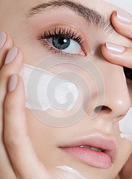 Young woman applying face cream or facial mask at her face. Beauty model with perfect fresh skin and long eyelashes cares about