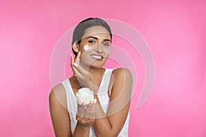 Young woman applying facial cream on pink background