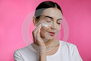 Young woman applying facial cream on pink background