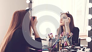 Young woman applying make-up while sitting at her vanity table with lots of cosmetics