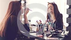 Young woman applying make-up while sitting at her vanity table with lots of cosmetics