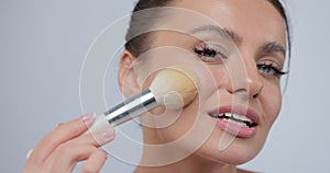 Young woman applying makeup blush with brush across her face while standing against studio gray background. Concept of