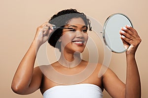 Young Woman Applying Mascara Holding Hand Mirror Against Beige Background