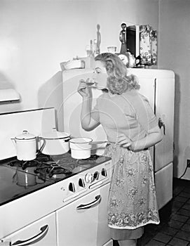 Young woman in an apron in her kitchen tasting her food from a pot